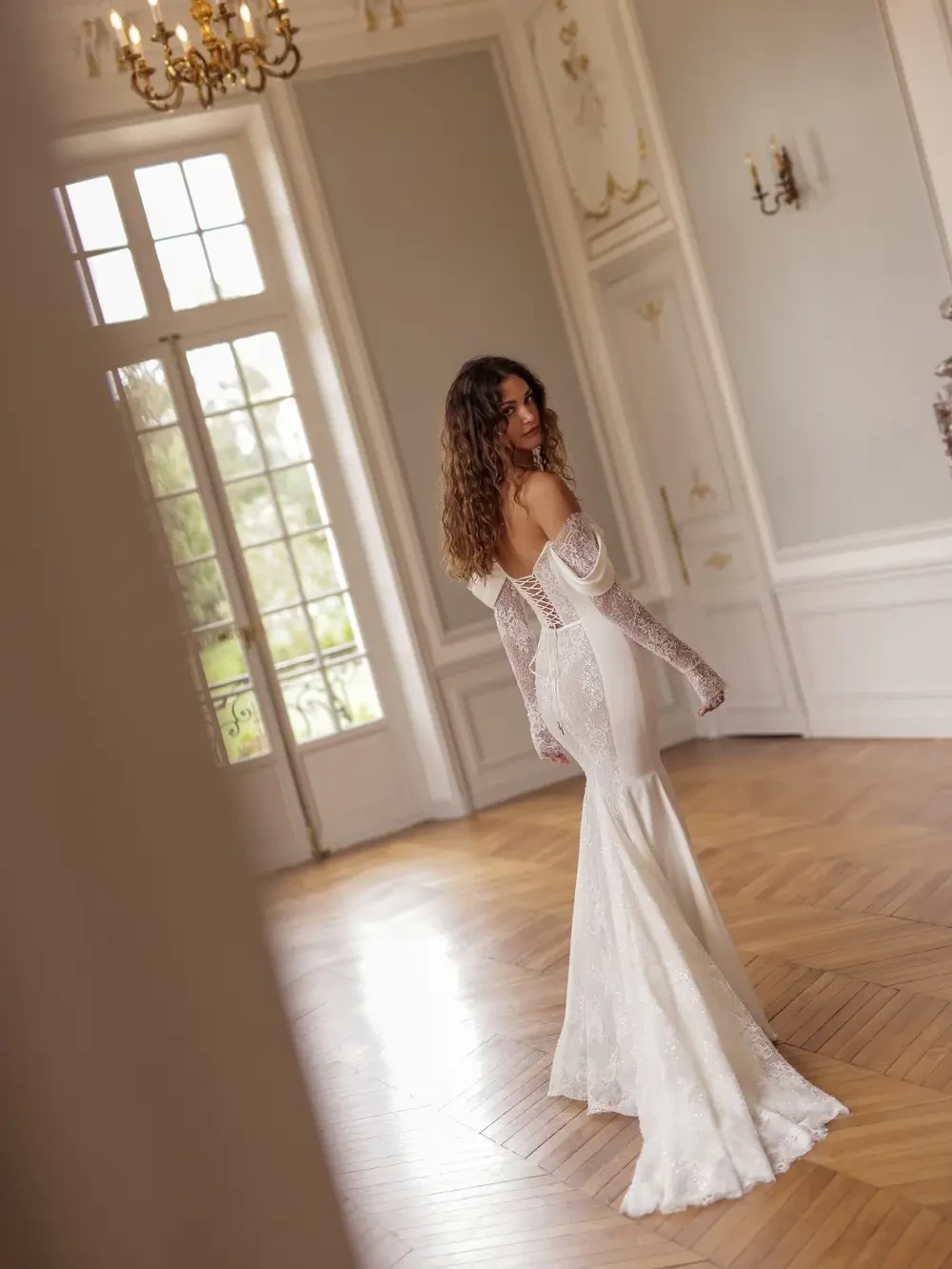A woman in an elegant white wedding dress with off-the-shoulder sleeves standing in a lavishly decorated room with ornate details and wooden flooring.