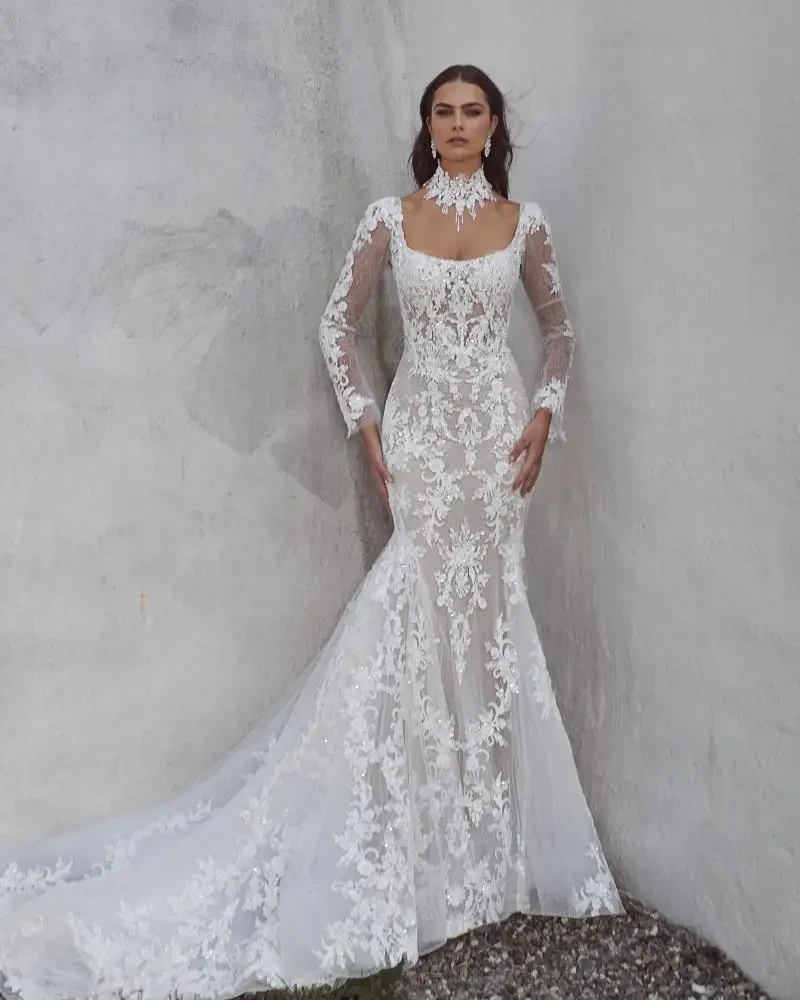 A model wearing a fitted white lace wedding dress with long sleeves and a train, standing against a textured wall.