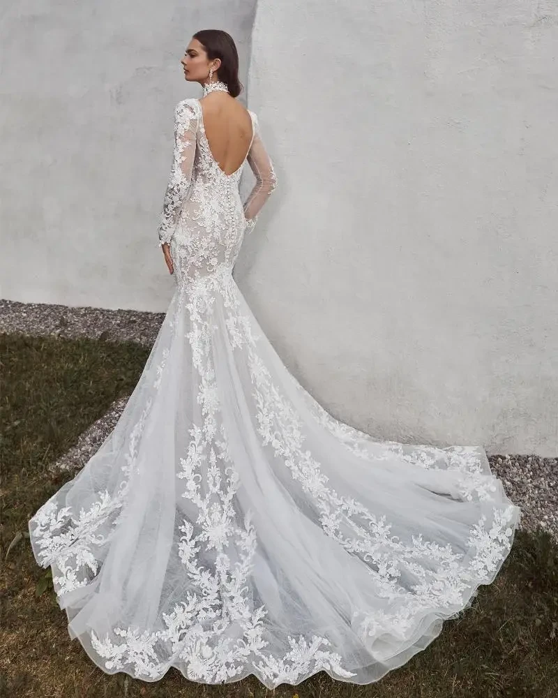 A model wearing a fitted white lace wedding dress with long sleeves and a train, standing against a textured wall.